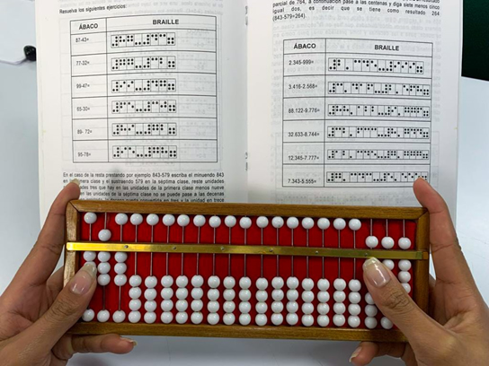 En primer plano aparecen unas manos utilizando un ábaco, enfrente hay una cartilla con operaciones matemáticas escritas en tinta y braille.