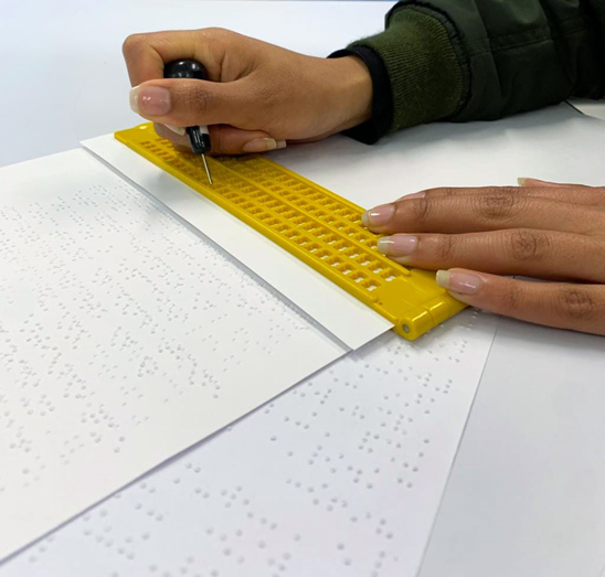 En primer plano aparecen unas manos escribiendo braille con pizarra y punzón, en frente reposan varias hojas ya escritas en braille.